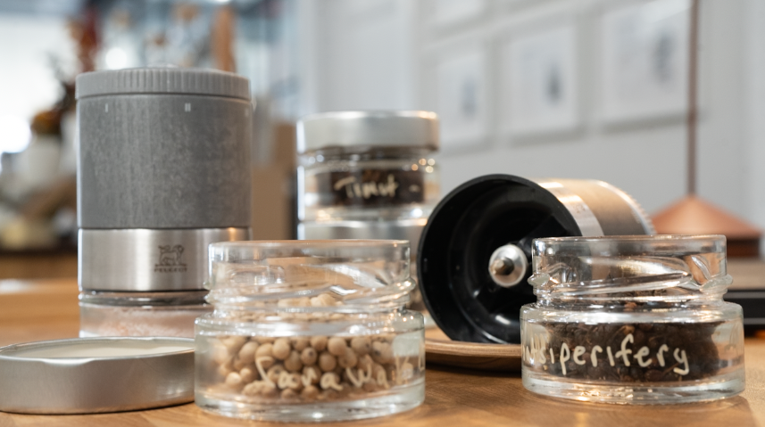 Collection of glass jars with metal lids on a wooden surface, likely used for storing spices or herbs with pepper and salt grinders. 